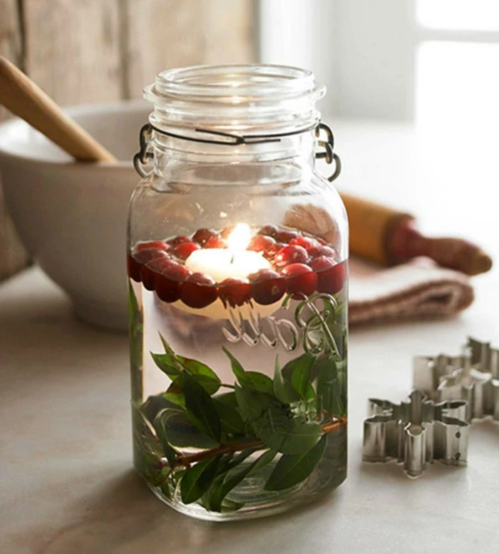 diy mason jar, filled with water, with small twigs with green leaves, cranberries and a lit candle swimming inside