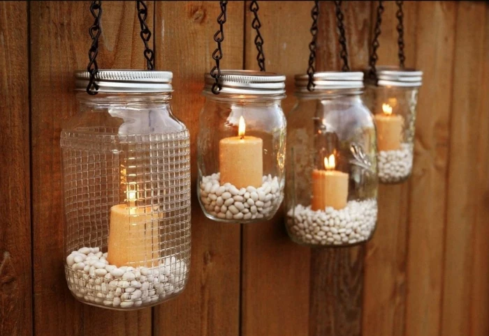 mason jar crafts, four jars with aluminium lids, half-filled with white beans, each containing a lit candle, hanging on black chains, from wooden wall