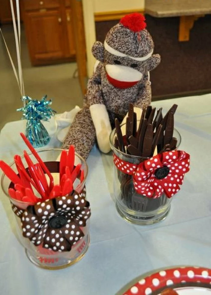 two small jars or glasses, decorated with brown or red ribbon, with bow and polka dots, containing red or brown plastic cutlery, placed on a table, near a stuffed monkey toy