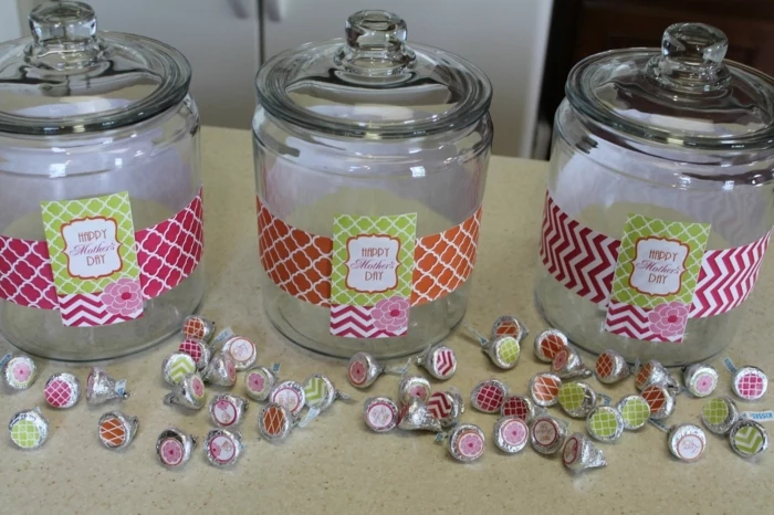 three empty candy jars, with glass lids, decorated with patterned labels in pink and green, orange and white, with many candies, in similarly colored wrappers nearby
