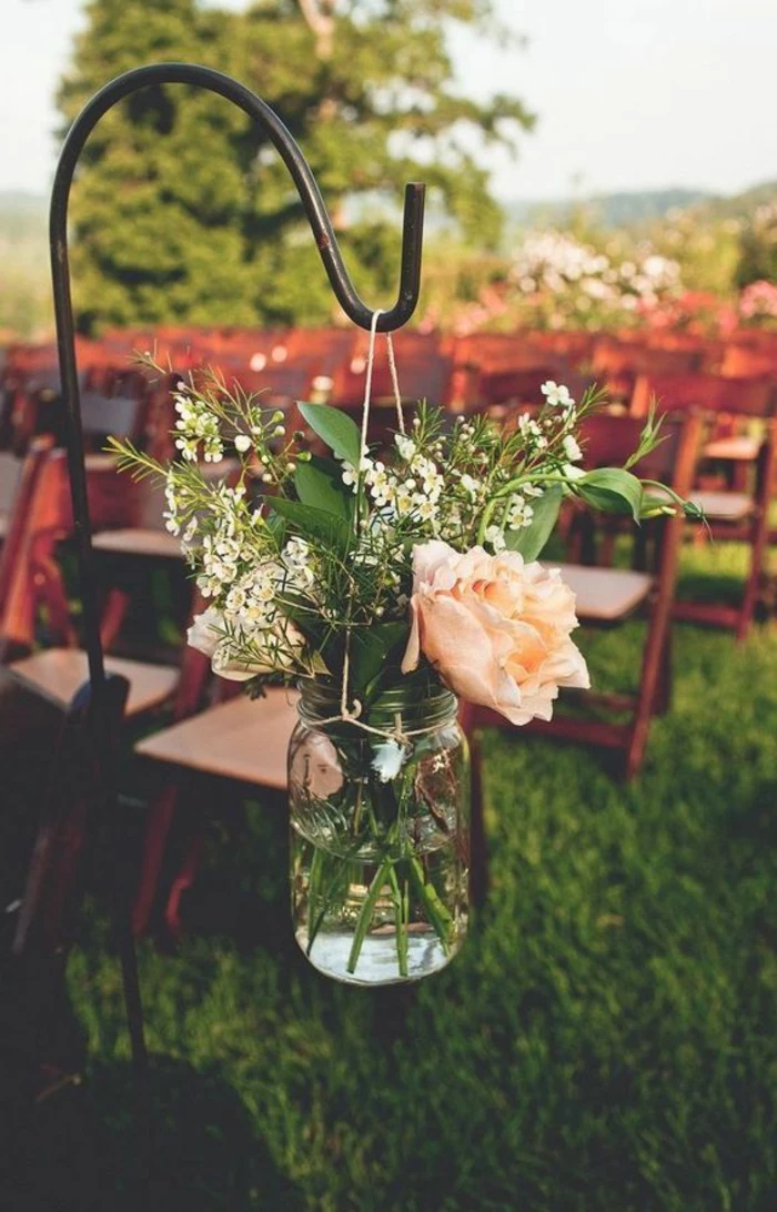 clear jar with string handle, hanging from a metal pole, containing a single pale pink rose, leafy green plants and tiny white flowers, rows of chairs on a meadow in the background