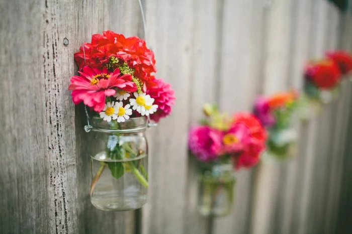 mason jar decorations, clear mason jars with wire handles, hanging from a wooden fence, containing daisies and red flowers