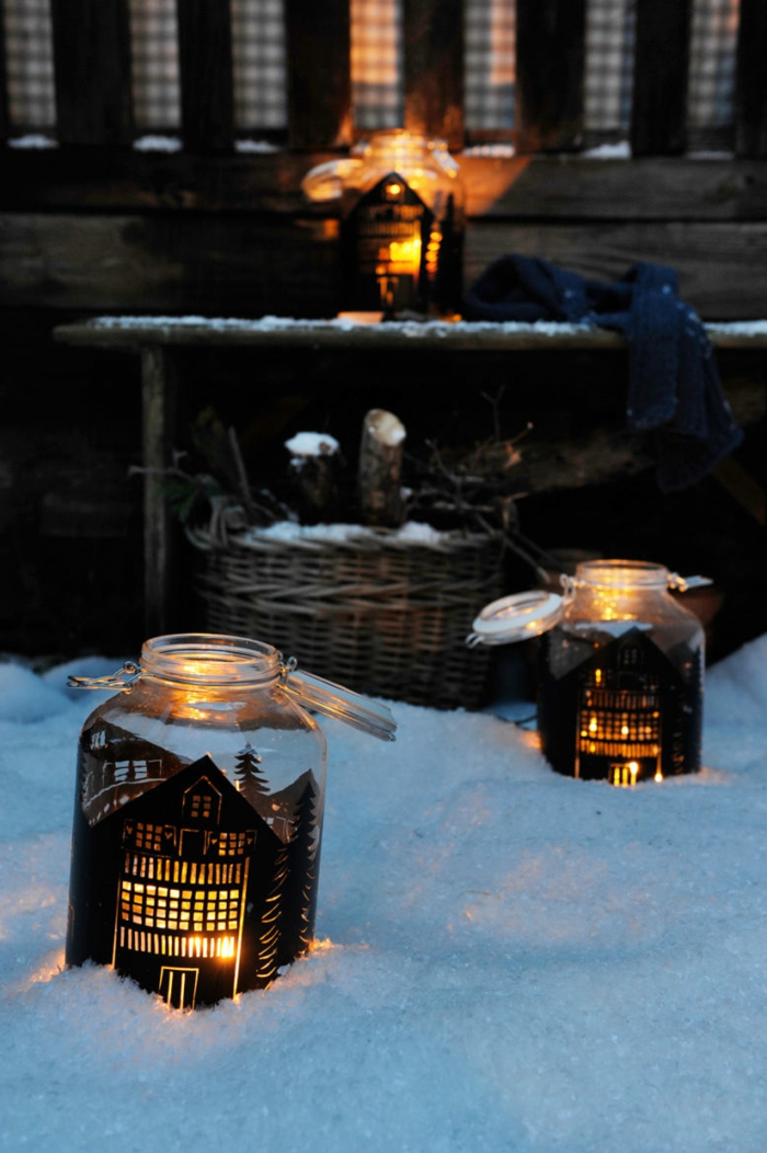 christmas mason jars, two jars with little houses painted on them in black, containing lit candles, and placed on snow near a house
