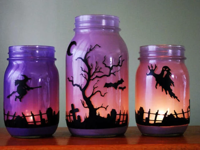 mason jar decorations, one large and two small jars, with lit candles inside, painted to look like an evening sky, with dark silhouettes of a witch, a ghost and a graveyard 