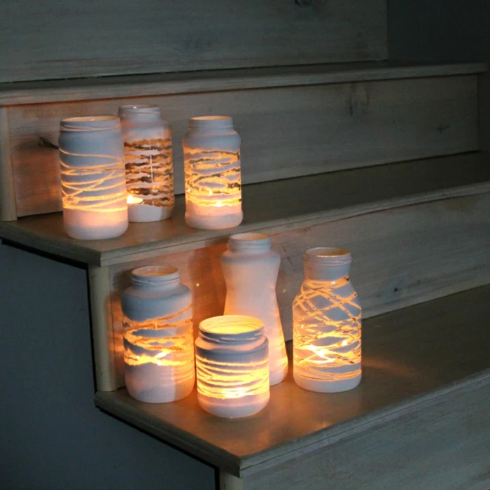 mason jar crafts, seven jars with different shapes and sizes, decorated with white paint in various patterns, and containing lit candles, placed on wooden steps 