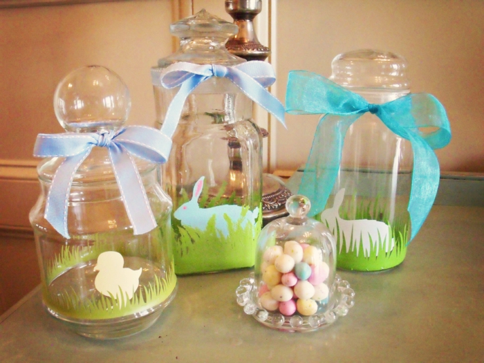 diy mason jar, three jars with clear lids, decorated with painted grass, bunnies and a chick, with blue and turquoise ribbons tied around the lids, and a small glass container with colorful candy nearby