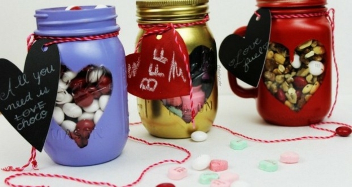 mason jar gifts, three jars painted in violet, gold and red paint, with heart-shaped windows, revealing colorful candy and snacks inside, heart-shaped labels in black and red, tied around them with string