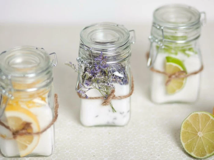 decorating mason jars, three clear jars with snap glass lids, containing hand-made candles, and decorated either with orange or lime slices, or tiny violet flowers, tied around each jar with string
