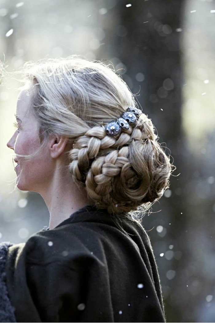 medieval braids, blonde braided hair, forming a voluminous bun at the back, decorated with silver ornaments, worn by woman in dark grey mantle