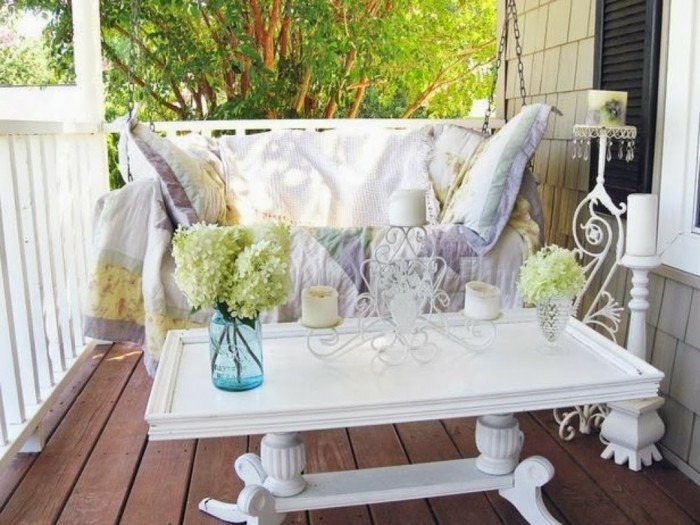 outdoor-patio-ideas-ornate-white-rectangular-table-with-candle-holder-blue-jar-and-crystal-vase-pale-green-hydrangeas-wooden-floor-and-settee-with-pale-cover-and-pillows couch with throw and pillow in light, pastel colors, near ornate white rectangular table, with candle holder and vases, wooden floor and front porch décor