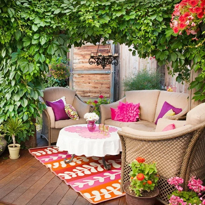 pale beige garden furniture, couch and two comfy seats, small round table, multicolored rug and potted plants, outdoor patio ideas, lots of greenery