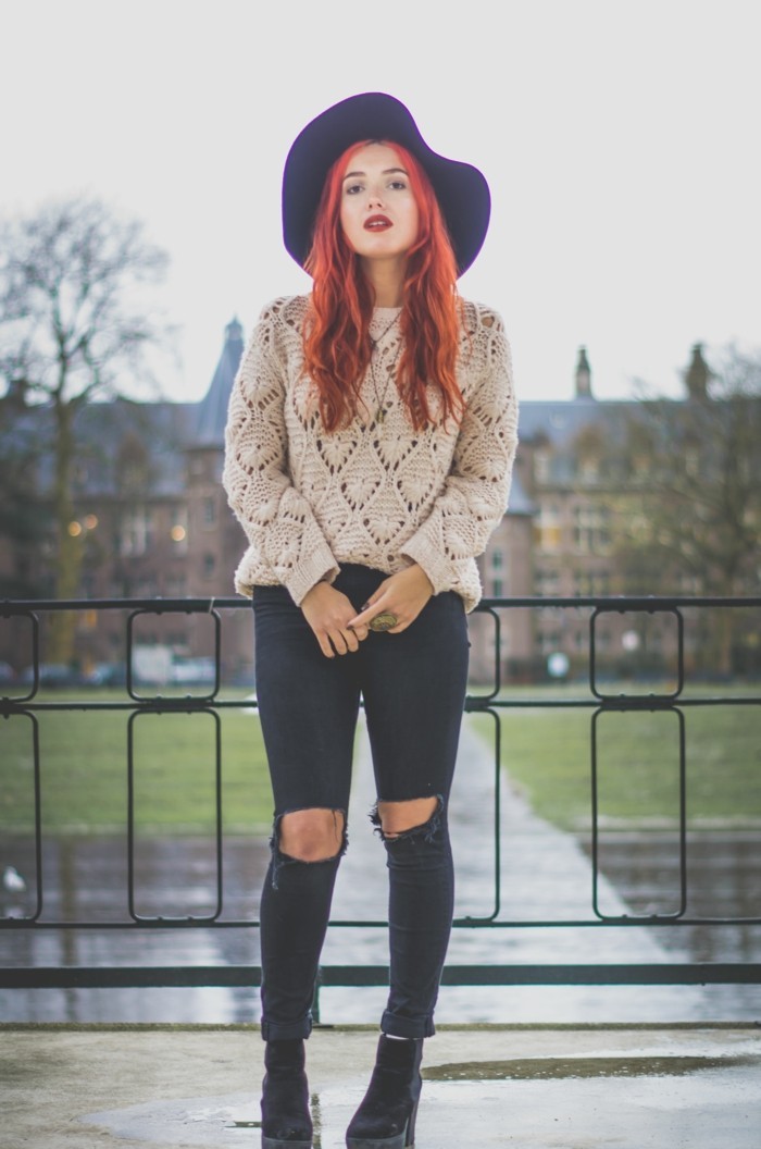 cute work outfits, young woman with long wavy red hair, wearing chinky knit pale beige jumper, and black torn skinny trousers, with dark ankle boots, and big felt hat