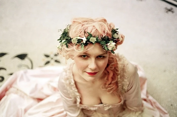 renaissance hairstyles, kirsten dunst, dressed as marie antoinette, with pale pink, frilly and shiny dress, pink curly hair in side ponytail, and green wreath with white flowers