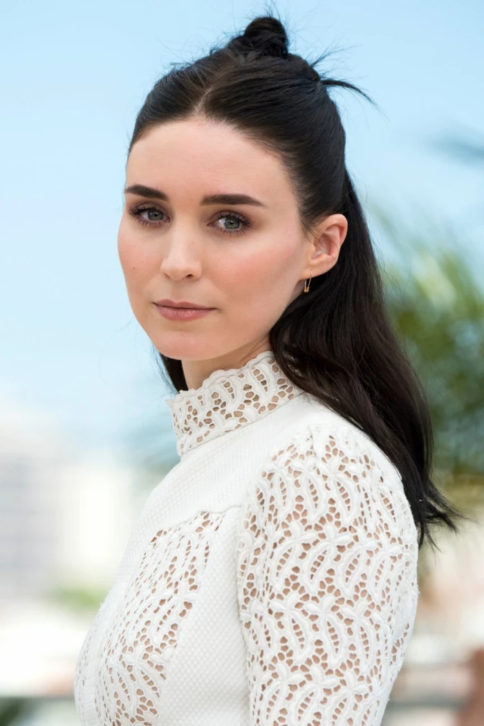 rooney mara, with dark hair parted in the middle, and small renaissance-inspired hair knot at the back, wearing white long-sleeved dress with lace details