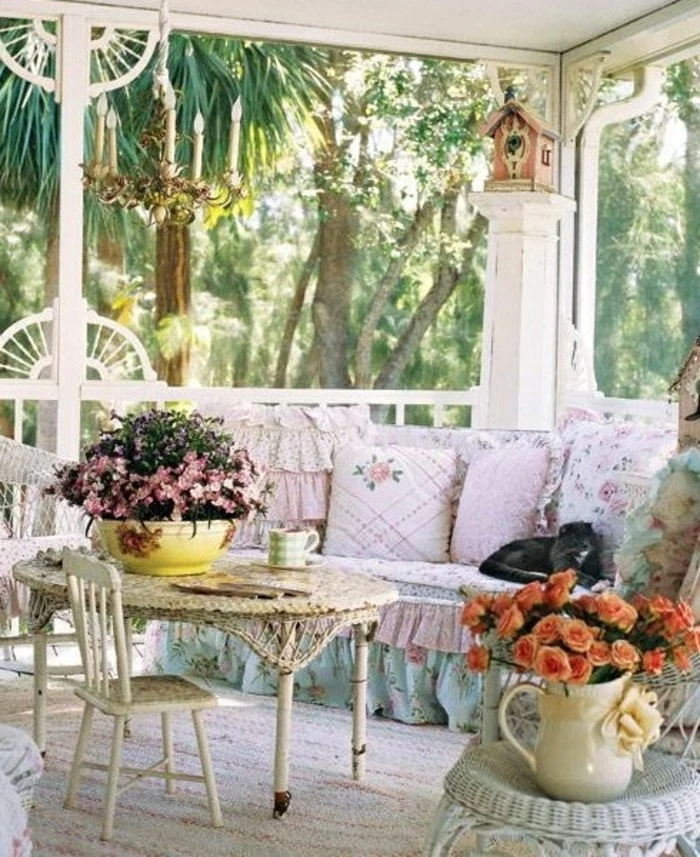 white round table with small chair, near couch with pale blue and pink cover, decorated with pale pink cushions, with frills and floral pattern, porch décor, many decorative objects