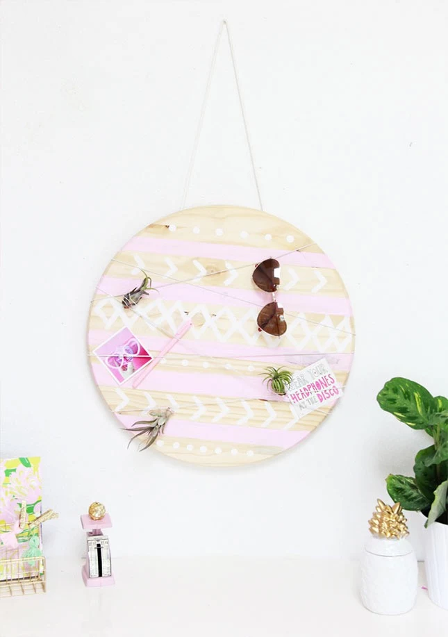 round wooden board on a wall, decorated with pale pink stripes, and white patterns, sunglasses and other items, hanging on it from a thread,