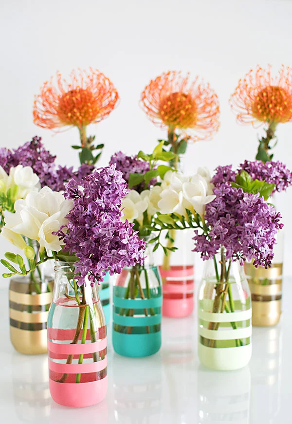 art and craft ideas, seven clear vases, partially painted with gold and pink, blue and white stripes, containing lilac stalks, white and orange flowers