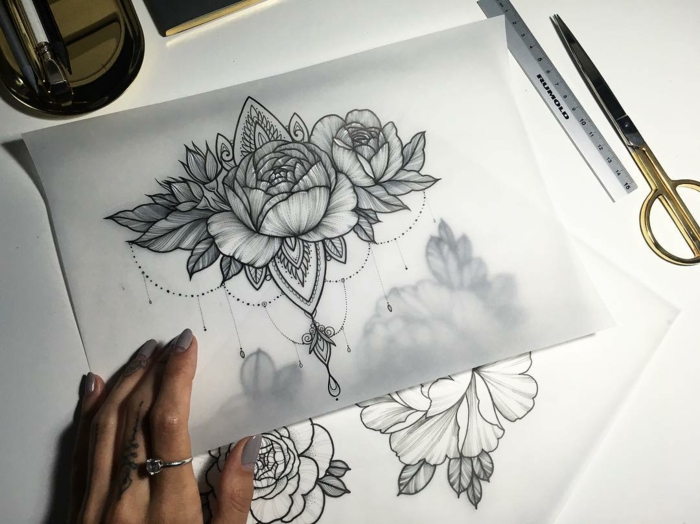 flower tattoo designs, hand holding sheet of tracing paper, with a drawing of a flower, and symmetric buddhist elements, more drawings underneath and stationary nearby