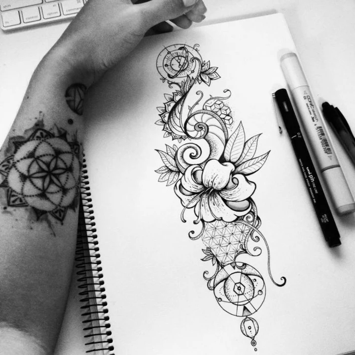 flower tattoo designs, male hand with mandala tattoo, leaning against a sketchbook, containing an ink drawing of a hibiscus flower, decorated with swirls and geometric patterns, fineliner pencil and marker nearby