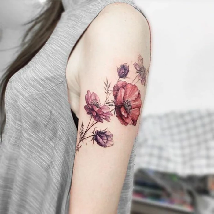 shoulder flower tattoos, brunette woman with long hair, and pale grey top, with a watercolor effect tattoo of several poppies, in pale red and violet, on her upper arm