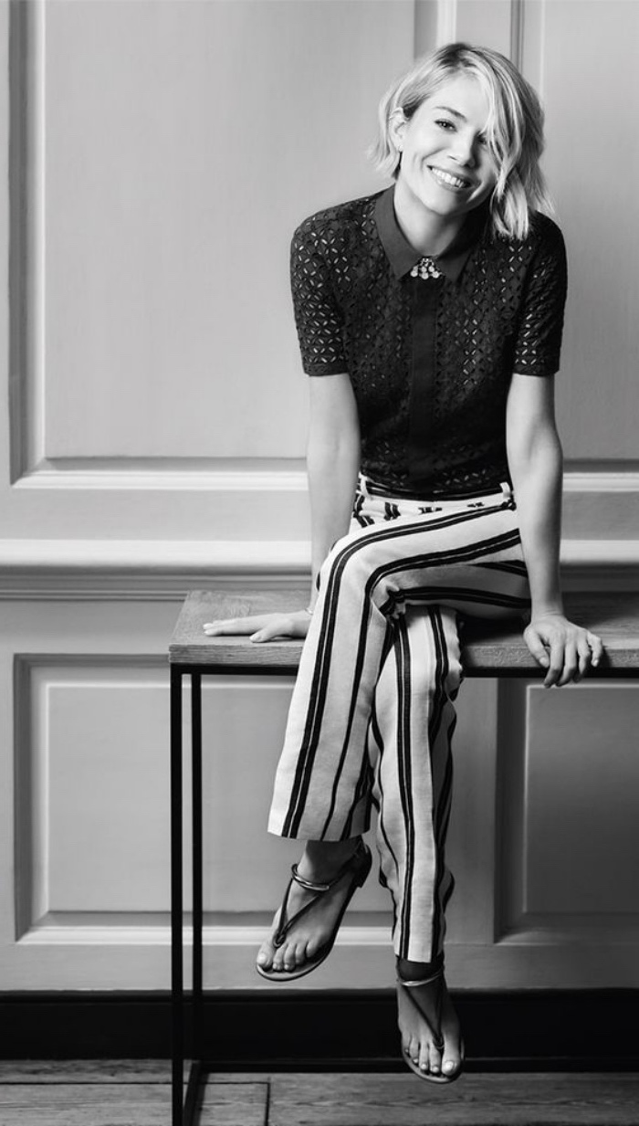 black and white photo of sienna miller, sitting on a table, wearing white pants with black stripes, black lace short-sleeved shirt, and a side parted, blonde wavy bob