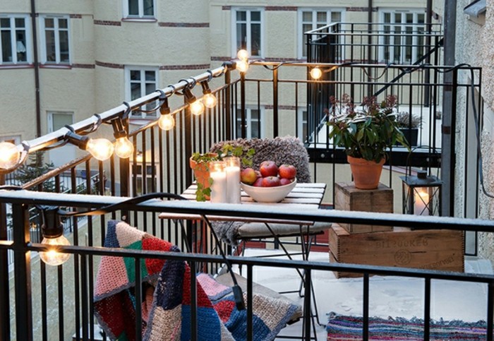 veranda with black railing, covered in a string of shining lightbulbs, small square wooden table with matching chairs, front porch decorating ideas, decorative wooden crates and potted plant