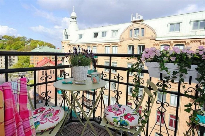 terrace with black ornate railings, with small round pale green table and matching chairs, decorated with cushions and pink blanket, potted plants and wooden laminate floor