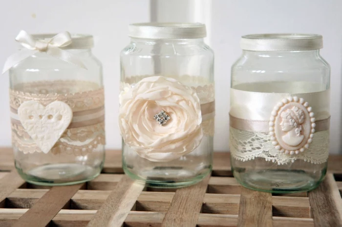 mason jar gifts, three clear jars, decorated with white lace and ribbon, one with a heart ornament, one with a faux flower, and one with a cream-colored brooch