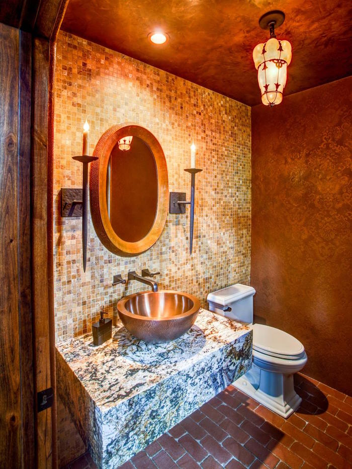 small bathroom ideas, toilet with brown walls, one with mosaic tiles, in different shades of brown, the other with brown paint and baroque pattern, large marble slab with metal sink, oval mirror with wooden frame, two lit candles in holders