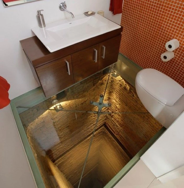 bathroom remodels, small toilet with two white walls, one wall covered by orange mosaic, brown wooden cupboard with flat white sink, white toilet seat, see through glass floor, revealing deep tunel beneath