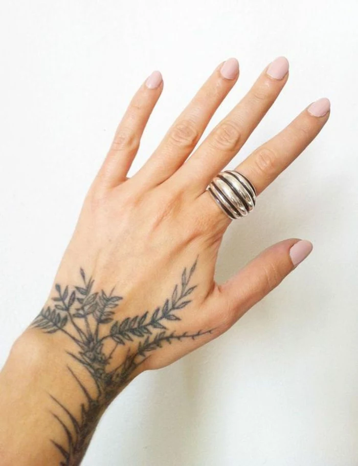 traditional flower tattoo, close up of a female hand, with pale pink nail polish, and large silver and black striped ring, a tattoo of a leafy plant going up the arm, and down towards the index finger