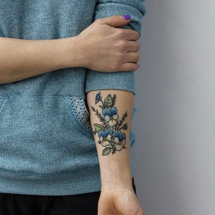 botanical tattoo, woman in pale blue sweater,with rolled up sleeves, holding her arm, with a tattoo of three pansies, in blue and yellow, with several buds and green leaves