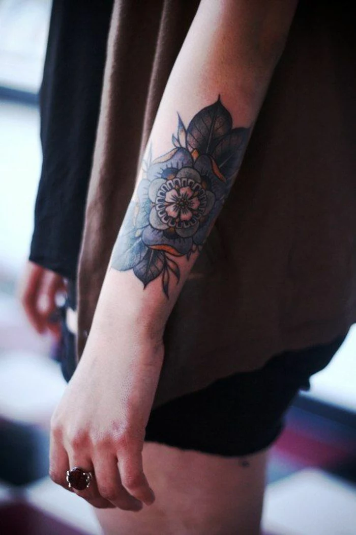 traditional flower tattoo, close up of a female lower arm, with an elaborate blue, green and yellow tattoo, with several rows of petals and green leaves, reminiscent of the tudor rose