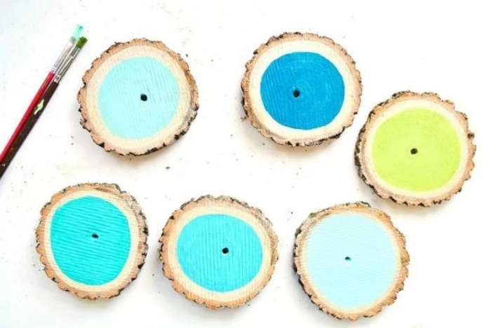 six round slices of wood, with bark, painted in different shades of blue and green, diy craft projects, paint brush nearby