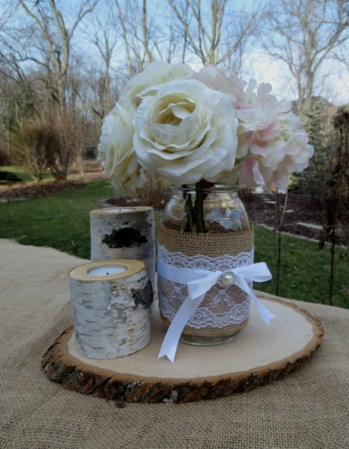 mason jar crafts, clear jar decorated with burlap, white lace and a ribbon with bow, containing white and pale pink flowers, candles nearby