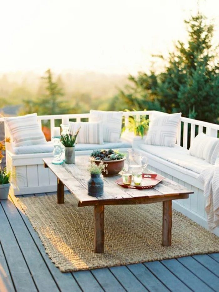massive wooden table, with vases and a trey with glasses and pitcher, white wooden corner settee, with pale pillows and blankets, beige coarse rug, outdoor patio ideas