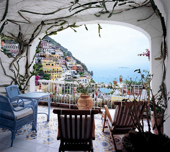 white-partly-covered-terrace-with-pale-blue-square-table-and-two-matching-chairs-two-wooden-lounge-chairs-with-white-covers-ornate-floor-tiles-cacti-on-walls-and-ceiling-beautiful-view pale blue chairs and table set, wooden lounging chairs, white walls and ceiling with ivy-like cacti, porch décor and beautiful view