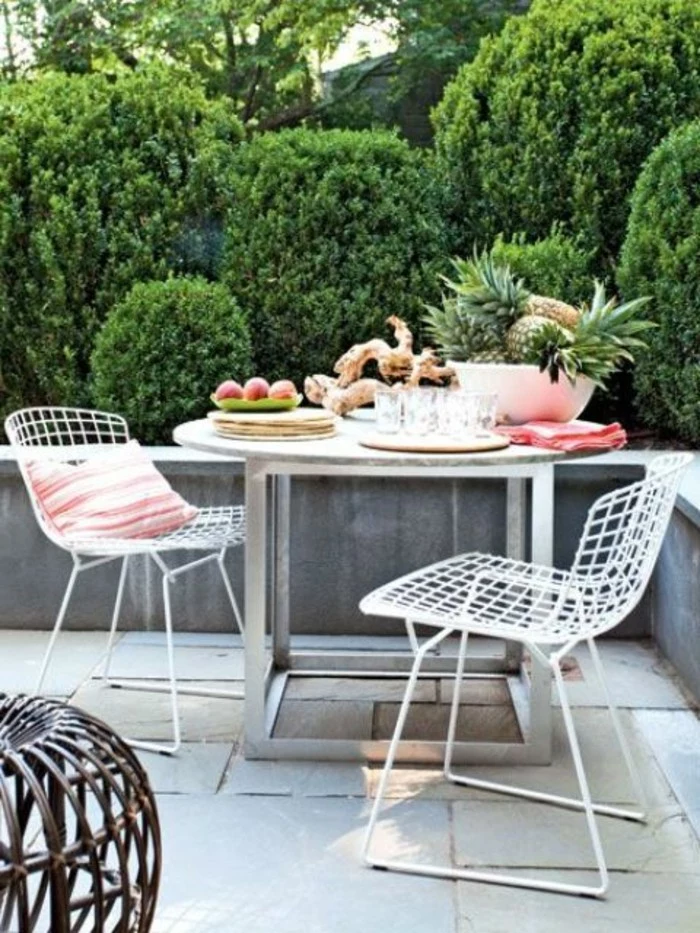 round metal table, with fruit and decorations, and two white chairs, one with pink striped pillow, outdoor patio ideas, greenery in background
