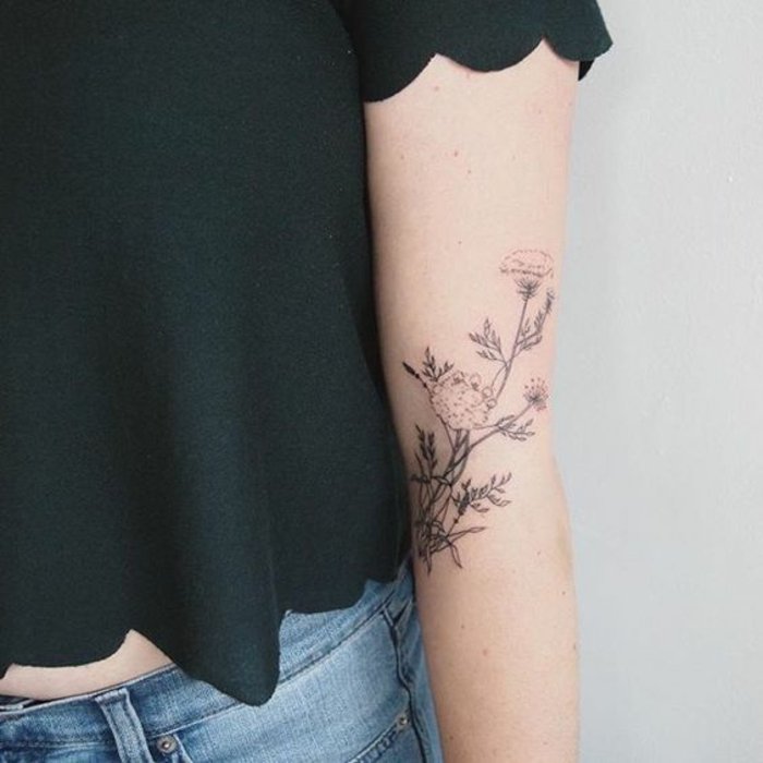 wildflower tattoo, woman with black cropped top, and blue jeans, with a tattoo of a wild flower, done with black ink, on her arm