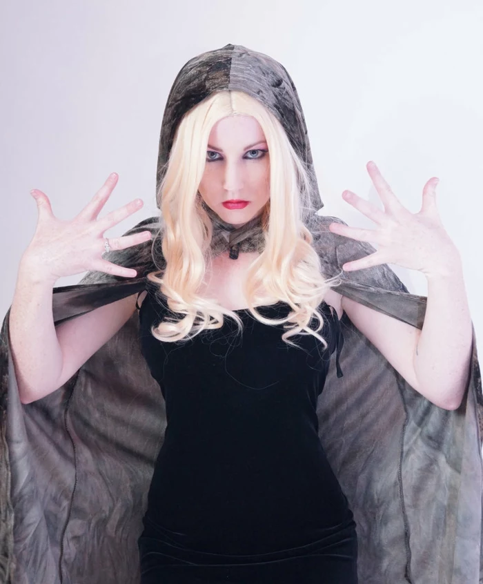 woman in blonde wig with curly ends, wearing black tight dress, and grey velvet hooded cape, heavy make up, and theatrical pose