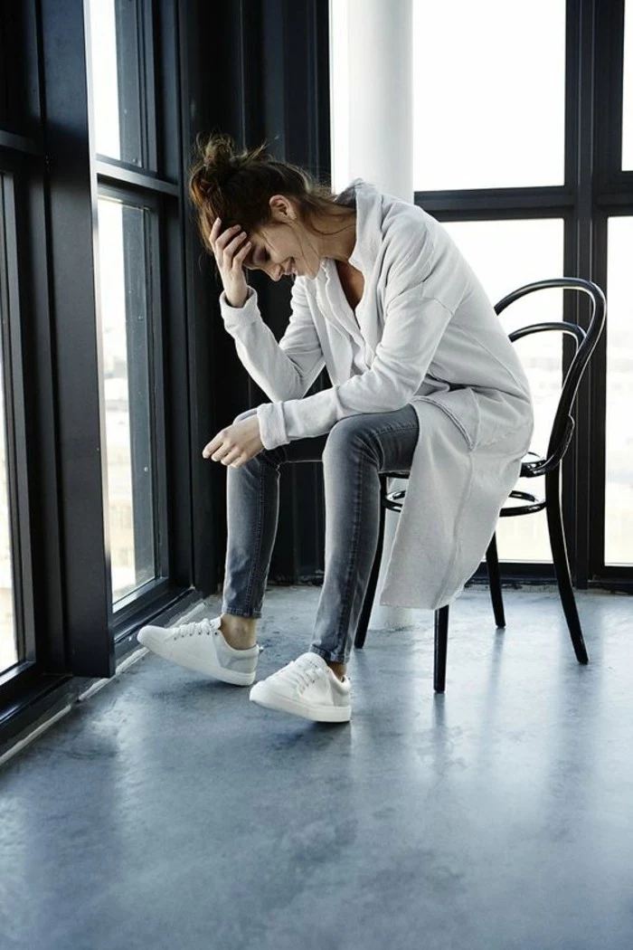 casual business attire, smiling brunette with messy top knot, sitting on a chair and holding her head with one hand, wearing grey skinny jeans, white sneakers and top, and long white cardigan