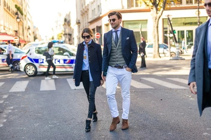 business casual for young women, brunette woman with pale blue shirt, dark jeans and navy blue coat, walking next to man in white trousers, with grey vest, pale shirt and dark tie