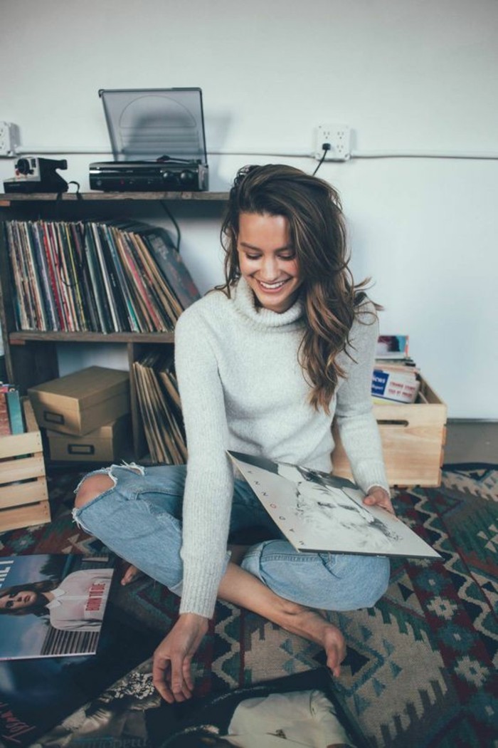 casual dress code, smiling brunette with wavy hair, wearing light grey turtleneck sweater, and pale ripped jeans with rolled legs, sitting on the floor among vinyl records