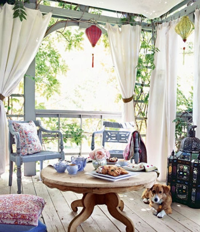 covered patio ideas, round wooden table, with tea set and pastries, two pale blue ornate chairs, covered patio ideas, white curtains and a dog