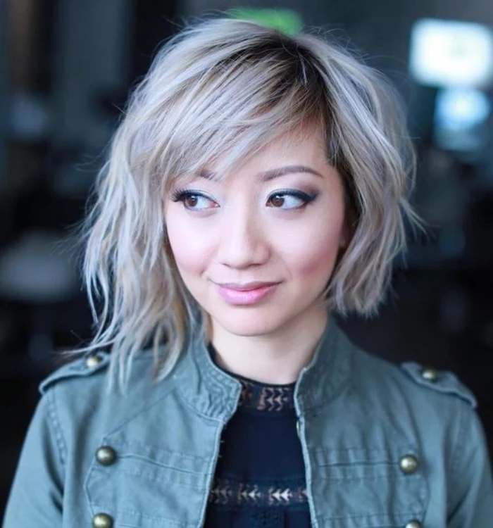 asymmetrical wavy hair, dyed platinum blonde, longer on one side, with side bangs and dark roots, worn by smiling woman, in pale green jacket, short bob hairstyles