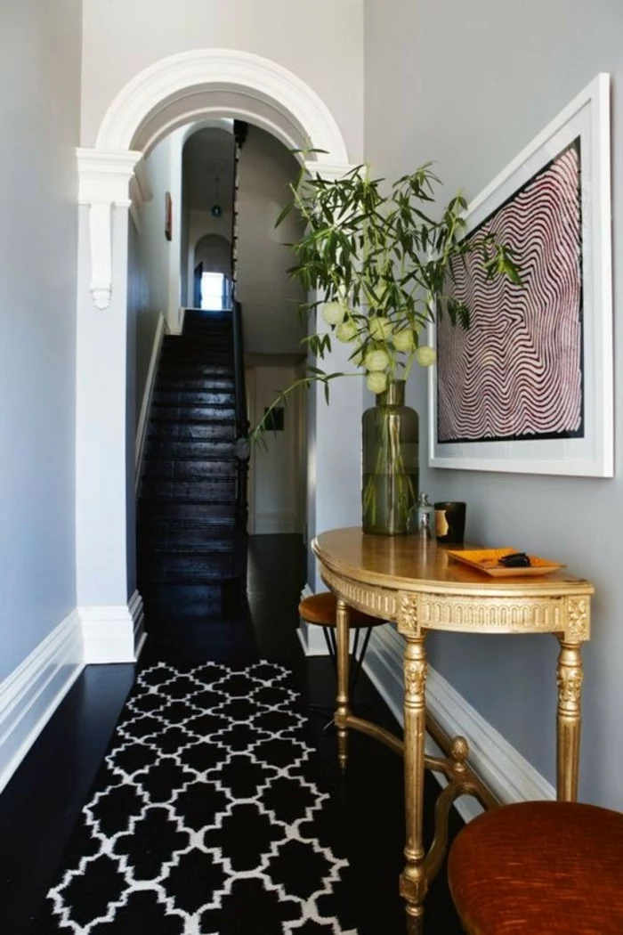 archway leading to a black staircase, black shiny floors, wooden table and two stools, framed abstract artwork, black and white patterned rug, long hallway runners