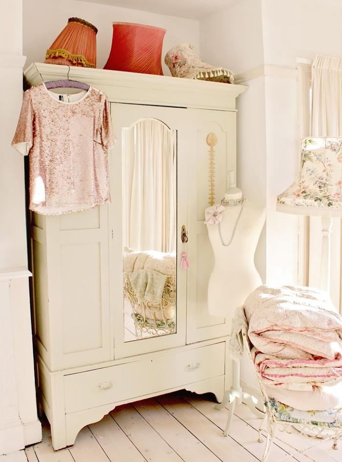 wardrobe in vintage style, painted white and featuring a mirror, inside a room with cream floors, shabby chic decorating, clothes and duvets on a vintage chair