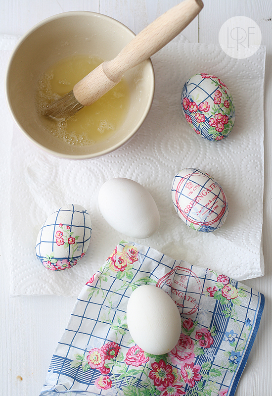 floral paper napkin in blue, pink and green, near two plain white eggs, and three eggs decorated with decoupage, coloring easter eggs, beige bowl with egg whites nearby