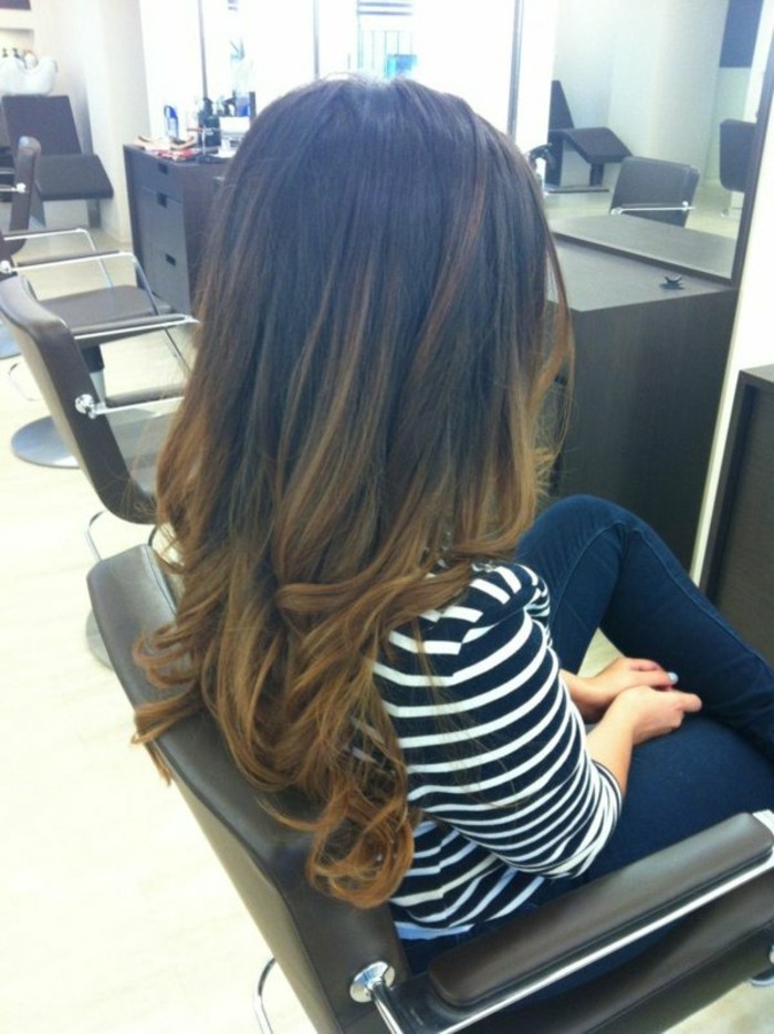 woman sitting on hairdresser's chair, with dark blue jeans, and white and black striped top, partially curled long, brown highlighted hair, with dark roots, and honey-blonde ends 