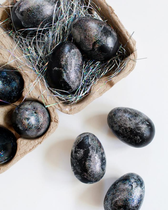 sparkly black eggs, covered in iridescent glitter, part of them are placed in a cardboard egg box, filled with hologram colored easter grass, others are placed on the table